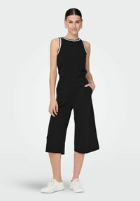 Svart byxdress med en figurnära överdel och vida cropped byxor. Har vitt detaljarbete längs halsringningen och sidofickor. Bärs med vita sneakers.