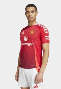 Maillot de football rouge à manches courtes avec une finition texturée, arborant un logo de club, un logo de sponsor et des accents blancs. Associé à un short blanc.