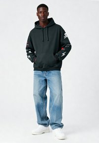 Felpa nera con tasca frontale, caratterizzata da vari design ricamati in rosso e bianco sulle maniche. Portata con jeans baggy azzurri chiari e sneakers bianche.