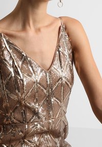 Donna che indossa un vestito senza maniche color oro rosa con paillettes e motivi geometrici e un piccolo orecchino a cerchio su uno sfondo chiaro.