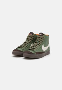 Zapatillas altas verdes con puntera de ante marrón, swoosh blanco de Nike y acento naranja en el collar; cuentan con cordones texturizados y lengüeta acolchada.