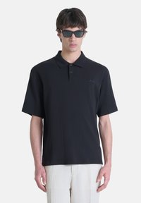 Crna pamučna polo majica s ovratnikom i tri dugmeta, s izvezenim logotipom na lijevoj strani prsiju. Nosena uz svijetle hlače.