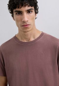 Støvet rosa T-shirt med korte ærmer, rund hals, lavet af blødt bomuldsstof, med et lille broderet logo på brystet. Glat tekstur.