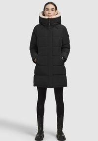 Frau in einem langen schwarzen gefütterten Mantel mit hohem Kragen und beigem Fleece-Futter, schwarzen Leggings und schwarzen Stiefeletten, die aufrecht steht.