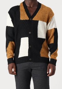 Kolorowy sweter na guziki o wzorze patchworkowym, łączący czerń, biel i odcienie brązu. Posiada dekolt w serek, guziki oraz ściągacze przy rękawach.