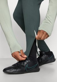 Tætsiddende mørkegrønne leggings med en sidelukning, parret med sorte sneakers med et teal Nike-logo. Lysegrøn langærmet top.