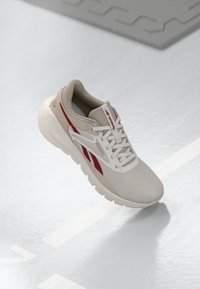 Zapato deportivo beige con acentos rojos, parte superior de malla texturizada, cuello acolchado y suela ligera. Presenta un logo en el lateral y un diseño con cordones.