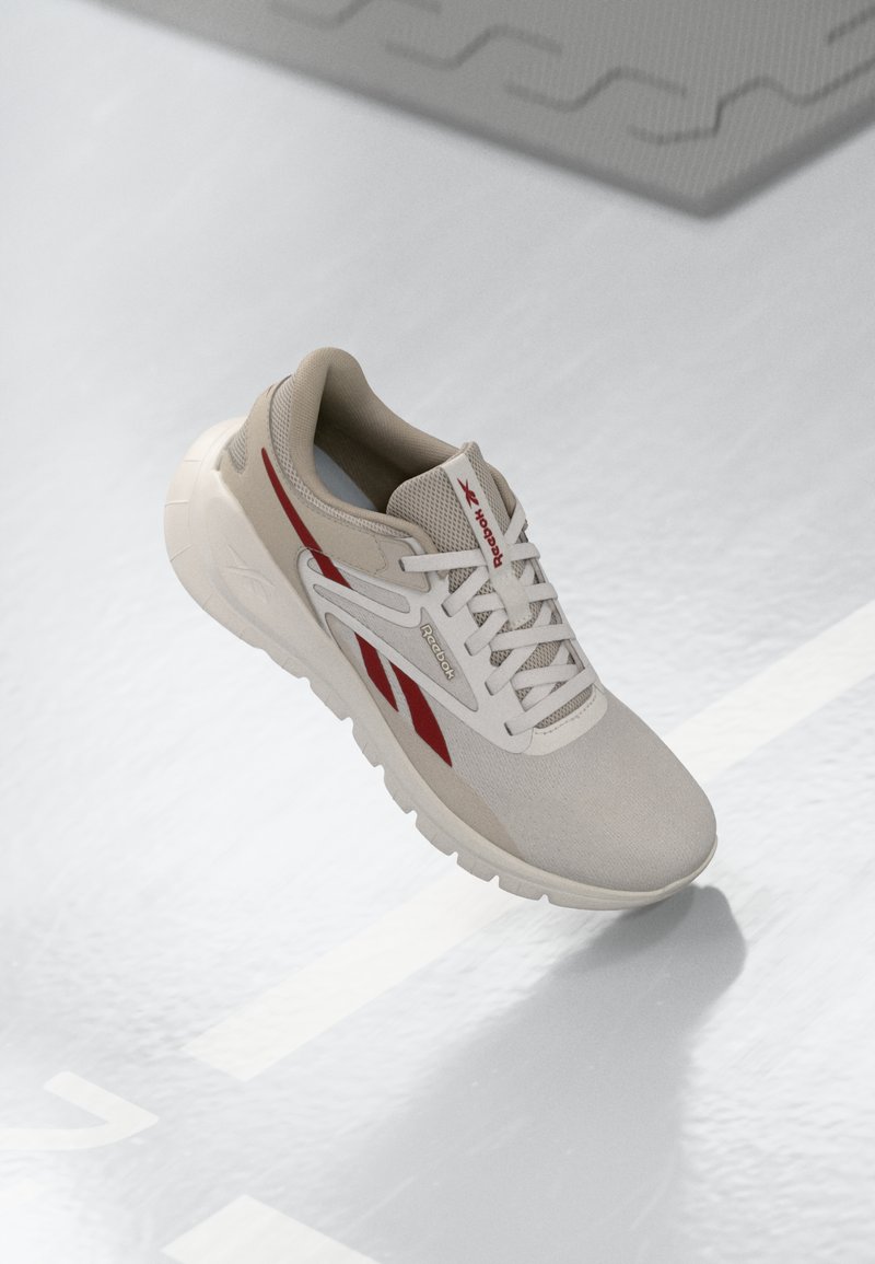 Zapato deportivo beige con acentos rojos, parte superior de malla texturizada, cuello acolchado y suela ligera. Presenta un logo en el lateral y un diseño con cordones.