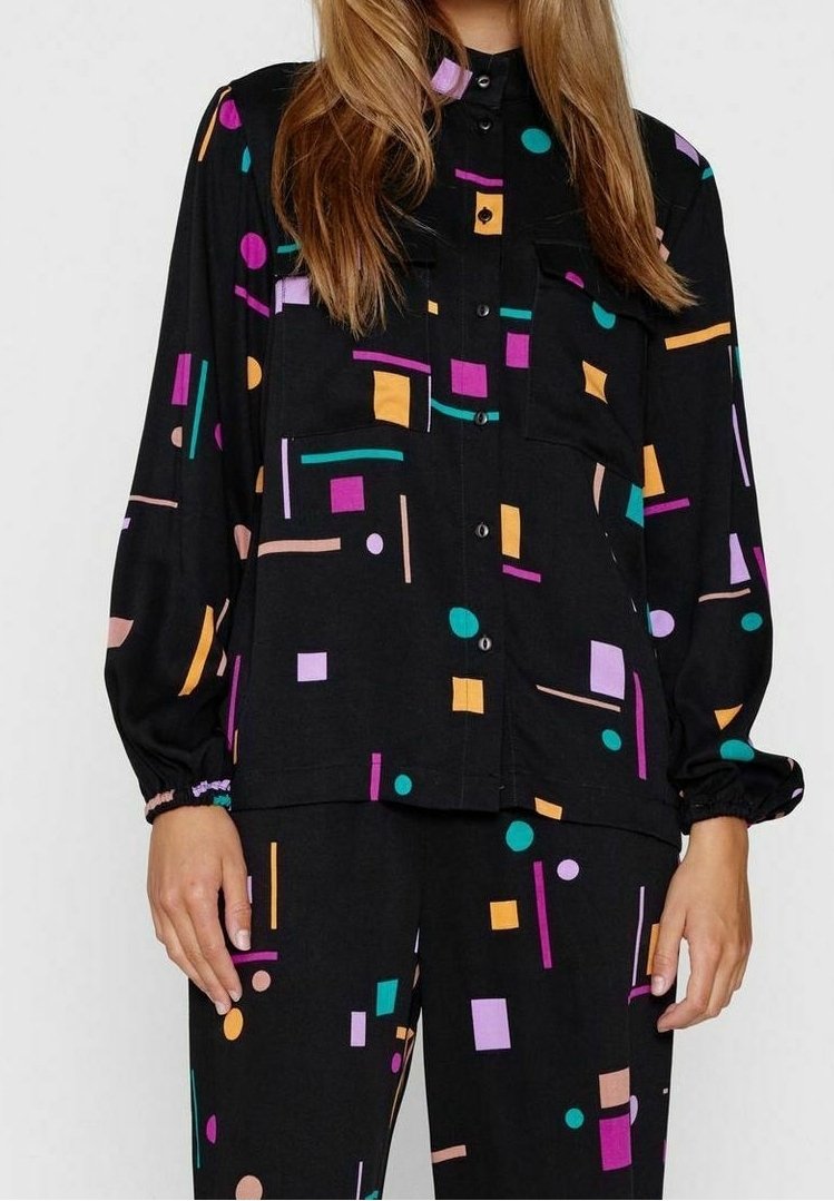 Femme portant une chemise noire à boutons et un pantalon avec des formes géométriques colorées en sarcelle, violet, orange et rose.