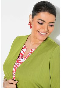 Zelená strukturovaná bunda přes červenou a bílou květinovou blůzu; velké červené květinové náušnice. Hladká pleť a neutrální pozadí.