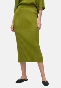 Jupe crayon en maille de couleur vert olive, avec des côtes verticales et une taille élevée. Portée avec des chaussures noires à enfiler.