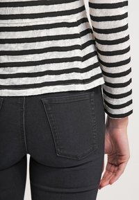 Personne portant un haut à manches longues rayé noir et blanc et un jean noir ajusté, montrant le dos avec la main droite détendue le long du corps.