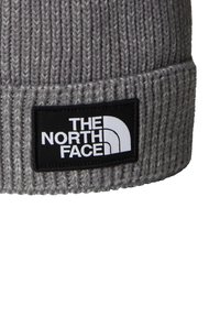 Šedý pletený čepice s hrubou texturou a ohrnutým lemem. Obsahuje černou obdélníkovou nášivku s bílým logem.