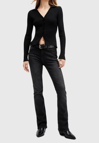Femme portant un cardigan noir côtelé boutonné, ceinture noire, jean noir délavé et bottes noires à bouts pointus sur un fond clair uni.