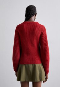 Femme aux cheveux tressés portant un pull rouge en tricot et une jupe plissée vert olive, debout face à un mur gris clair.