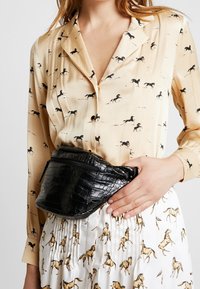 Donna che indossa una blusa beige e una gonna bianca con stampe di cavalli, che tiene una borsa a vita in pelle nera testurizzata in vita.