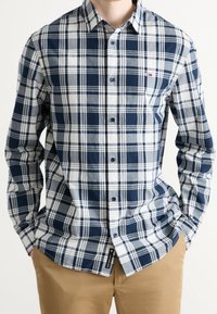 Mężczyzna w granatowo-białej i szarej koszuli w kratę z długim rękawem, noszący beżowe spodnie, z rękami w kieszeniach, na jednolitym tle.