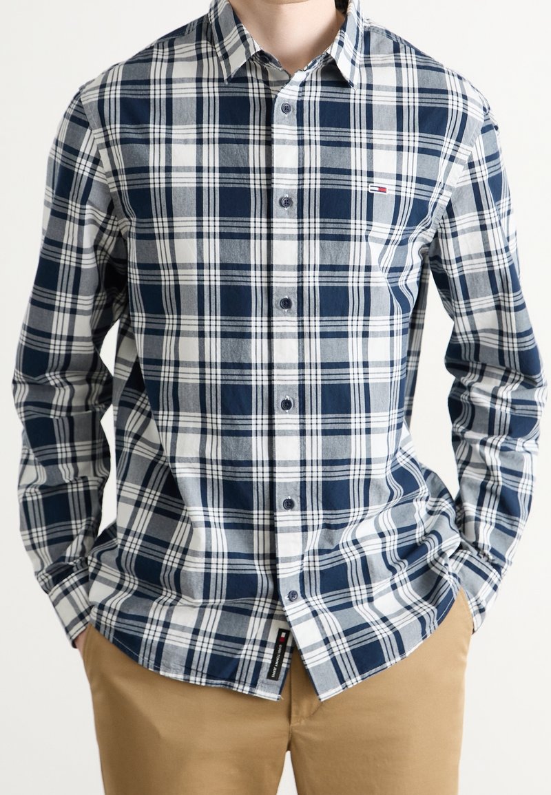 Mężczyzna w granatowo-białej i szarej koszuli w kratę z długim rękawem, noszący beżowe spodnie, z rękami w kieszeniach, na jednolitym tle.