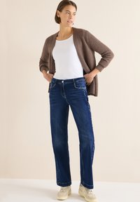 Vaqueros de mezclilla azul con pierna recta, costuras laterales detalladas y cierre de botones, combinados con una camiseta sin mangas blanca y un cárdigan marrón.