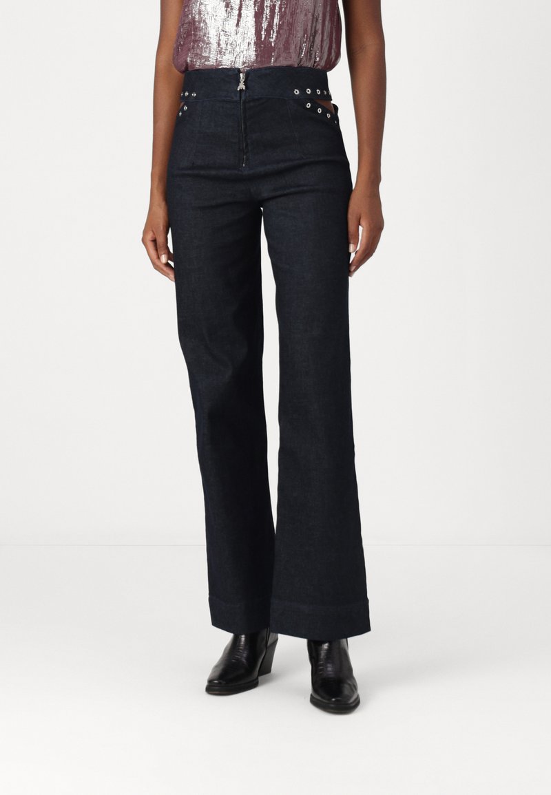 Pantalones de mezclilla en azul oscuro con cintura alta, cremallera frontal y ojales decorativos en plata. Piernas amplias con un detalle de dobladillo enrollado.
