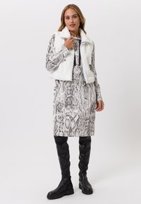 Witte cropped faux fur-vest over een jurk met slangenhuidpatroon; kniehoge zwarte leren laarzen; lange mouwen met een smalle pasvorm.