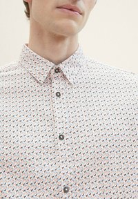 Korte mouwen overhemd in het wit met een geometrisch patroon met oranje, blauwe en grijze vormen. Knopsluiting aan de voorkant met donkere knoppen.