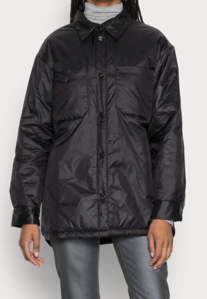 Personne portant une veste noire brillante à boutons avec poches poitrine, un col roulé gris et un pantalon gris foncé sur un fond uni.