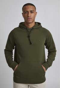 Sudadera con capucha color verde oliva, con un bolsillo frontal, cierre con cordón y puños acanalados. Presenta un pequeño logo en el pecho.