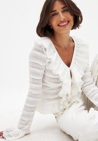 Blouse blanche à manches longues avec un col et des poignets à volants. Présente un motif texturé à rayures avec des sections transparentes et un décolleté en V.