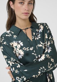 Grünblumenbluse mit langen Ärmeln, Kragen und einer Bindedetail am Hals. Verfügt über cremefarbene und hellblaue Blumenmuster auf satiniertem Finish.