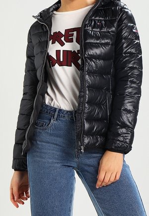 Personne portant une doudoune noire par-dessus un T-shirt blanc avec un motif graphique en rouge et un jean bleu taille haute, debout avec une main détendue.