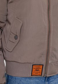 Giubbotto da volo marrone con zip frontale, orlo a costine e una tasca a sinistra. Presenta un'etichetta arancione con testo nero. Tessuto dalla consistenza liscia.