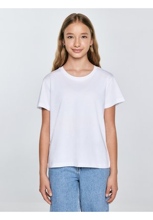 Jauna meitene ar gariem blondiem matiem, tērpusies vienkāršā baltā T-kreklā un gaiši zilās džinsās, stāvot pret gaiši pelēku fonu.