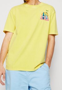 Camiseta de algodón amarilla con un corte relajado. Presenta gráficos bordados coloridos de bebidas y palmeras en el lado izquierdo del pecho.