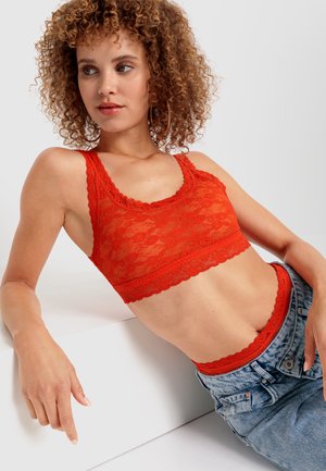 Bralette de encaje rojo con bordes festoneados, combinado con pantalones cortos de mezclilla. El encaje presenta patrones florales, exhibiendo una textura transparente.