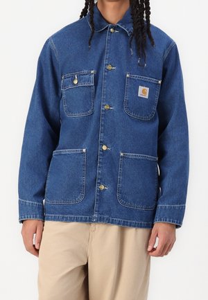 Mann trägt eine blaue Jeansjacke mit Knöpfen und vier vorderen Taschen, davon zwei Brusttaschen, eine mit Carhartt-Logo-Patch, und beigefarbene Hose.