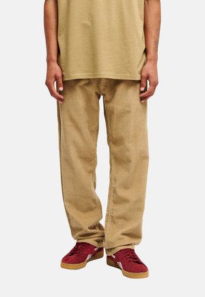 Personne portant un pantalon en velours côtelé beige, une chemise vert olive et des baskets en daim rouge avec semelles en gomme, debout devant un fond uni.