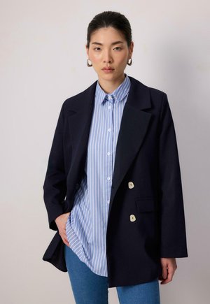 Námořnický dvouřadový blazer s oversized klopami, zkombinovaný s světle modrou pruhovanou košilí s knoflíky a dobře padnoucími modrými džíny.