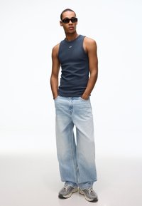 Marineblauw mouwloos shirt, lichtblauwe jeans met brede pijpen, grijze sneakers met zwarte accenten en donkere zonnebril. Minimalistisch ontwerp met lichte textuur.
