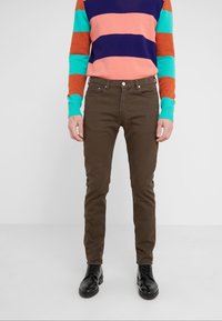 Hnědé slim-fit džíny vyrobené z bavlněné směsi, nošené s multibarevným pruhovaným svetrem. Černé šněrovací boty doplňují outfit.