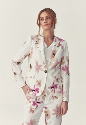 Kvinde iført hvidt jakkesæt med pink og beige blomsterprint, poserer med den ene hånd i lommen mod en ensfarvet, lys baggrund.