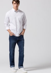 Camicia bianca a maniche lunghe con bottoni e tasca, abbinata a jeans blu scuro e sneakers bianche. Tessuto morbido, vestibilità comoda, polsini ripiegati.