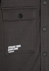Mørk grå denimskjorte med en brystlomme og en svart knapp. Teksten sier "UPGRADE YOUR DENIM GAME" i hvitt, med merkevarenavnet @VINGINO.