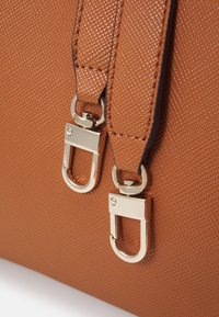 Hnědá texturovaná syntetická taška se stříbrnými kovovými přezkami a kováním. Má hladké šití a strukturovaný design.