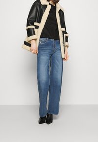 Cappotto di pelle nera con foderato in shearling beige e righe, abbinato a jeans a gamba larga blu chiaro e stivaletti neri con tacco.