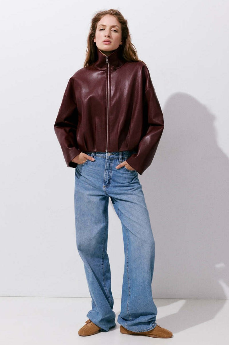 Femme debout, les mains dans les poches d’un jean large bleu clair, portant une veste en cuir marron foncé zippée et des chaussures marron.