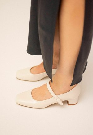 Scarpe Mary Jane color crema con punta quadrata, una sola cinghia alla caviglia e un tacco basso, esposte su uno sfondo chiaro.