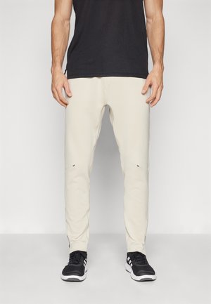 Mężczyzna ubrany w kremowe, dopasowane spodnie i czarne buty sportowe, stojący na tle jednolitej białej ściany.