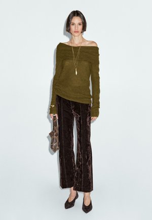 Olijfgroene off-shoulder trui, flared donkerbruine fluwelen broek en puntige hakken; geaccessoiresd met een slangenprint handtas en een gelaagde ketting.