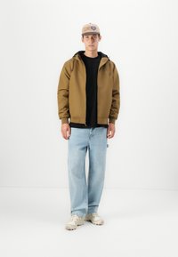 Bruine zip-up jas over een zwart shirt, gecombineerd met lichtblauwe losse jeans en beige sneakers. Een beige pet met een logo maakt de look compleet.